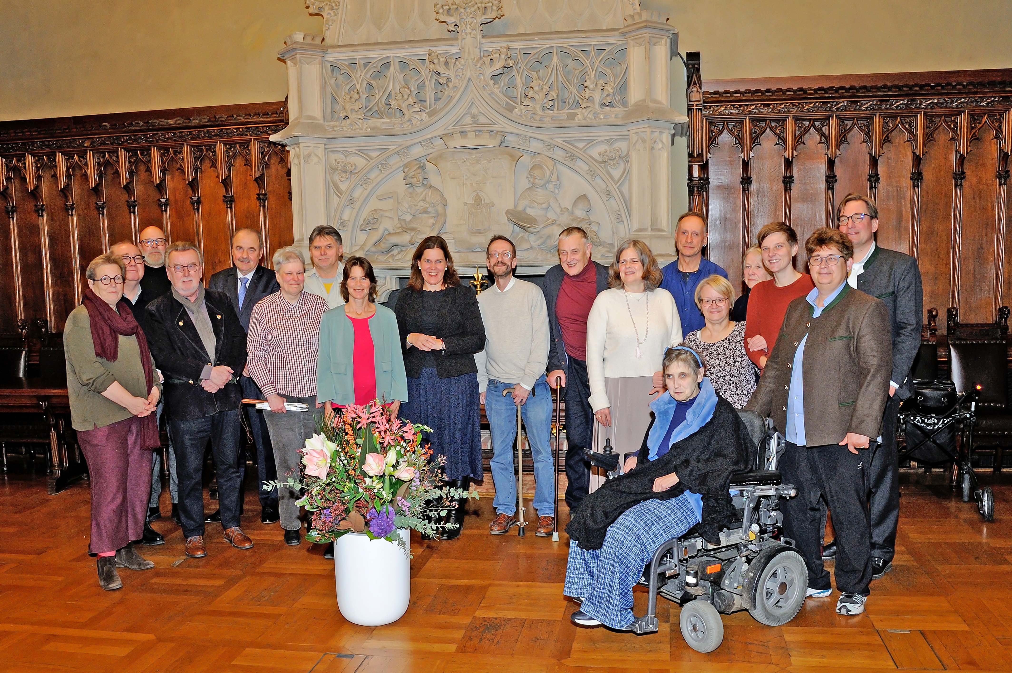 eine Gruppe von Menschen in einem historischen Raum mit verziertem Hintergrund und einem großen Blumenarrangement im Vordergrund.