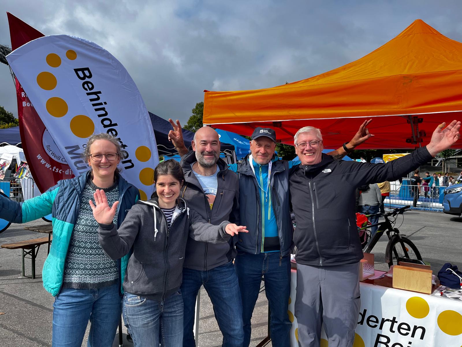 Fünf fröhliche Personen stehen vor einem Infostand beim Outdoor-Festival. Sie lachen in die Kamera und winken. Im Hintergrund sind ein orangefarbenes Zelt und eine Fahne mit der Aufschrift ‚Behinderten­beirat München‘ zu sehen.