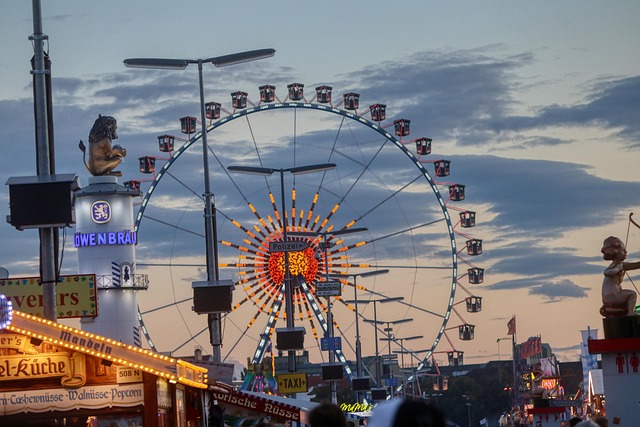 Abendaufnahme vom Oktoberfest in München mit beleuchtetem Riesenrad im Hintergrund, bunten Fahrgeschäften und Bierzeltschildern im Vordergrund.