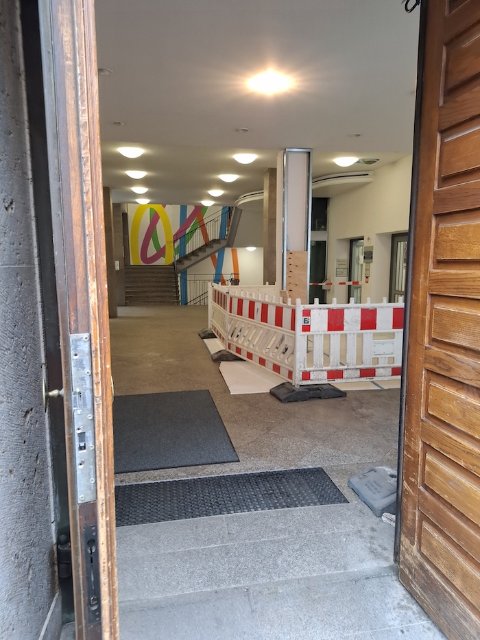 Blick durch die geöffneten Holztüren in den Eingangsbereich der Burgstrasse 4. Im Foyer ist ein Bereich wegen Bauarbeiten mit rot-weißen Absperrgittern gesichert. Im Hintergrund führen Treppen nach oben; die Wände sind mit einer großflächigen, farbenfrohen Gestaltung versehen.