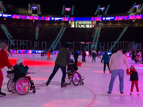Menschen nehmen gemeinsam an einem inklusiven Eislauf in einer großen Eishalle teil. Zwei Kinder sitzen in Rollstühlen und werden auf dem Eis von Erwachsenen begleitet. Weitere Teilnehmende bewegen sich auf Schlittschuhen. Bunte Beleuchtung und Anzeigetafeln sind im Hintergrund sichtbar.