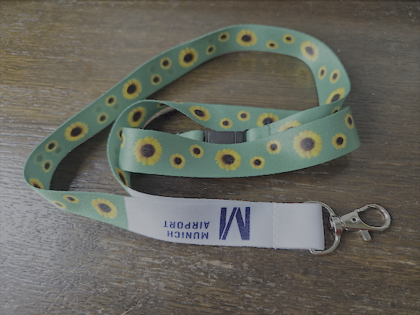 Grünes Schlüsselband mit Sonnenblumenmuster und Metallkarabiner liegt auf einer dunklen Holzoberfläche. Am weißen Bandende ist das blaue Logo „Munich Airport“ zu sehen.