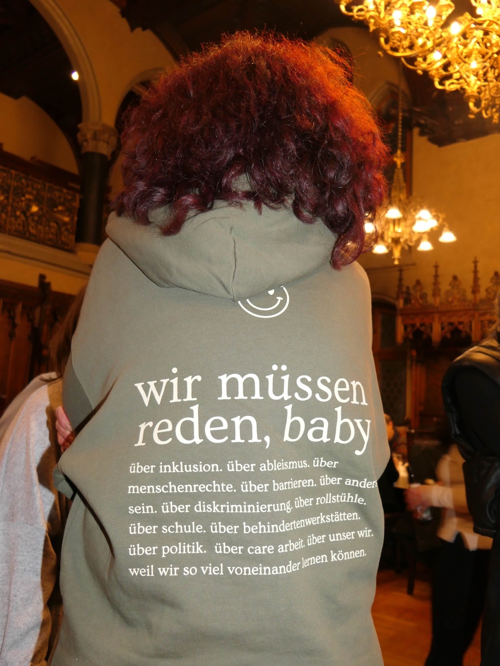Eine Person mit lockigem rötlichem Haar steht in einem festlich beleuchteten Raum und trägt einen Hoodie. Auf dem Rücken des Hoodies steht: „wir müssen reden, baby“ und darunter eine Liste von gesellschaftlichen Themen wie Inklusion, Ableismus, Menschenrechte, Barrieren, Diskriminierung, Schule und Politik.