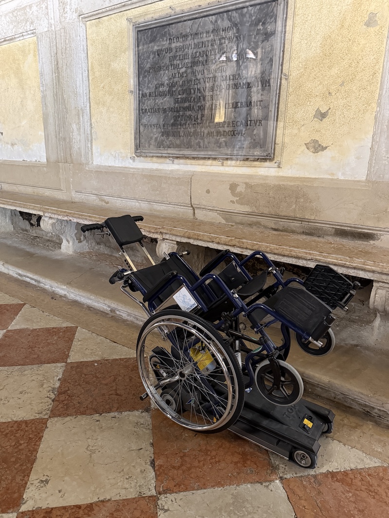 Zusammengeklappter Rollstuhl mit einer mobilen Treppensteighilfe auf einem Steinboden in einem historischen Gebäude in Venedig. Im Hintergrund befindet sich eine helle Wand mit einer eingelassenen Gedenktafel und sichtbaren Gebrauchsspuren.